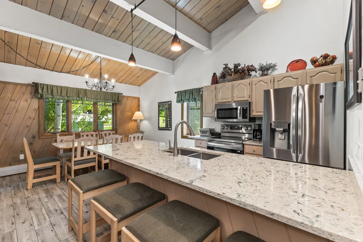 A warm, inviting kitchen and dining area with a granite island, wooden beams, and large windows. The dining table is set for a group, and the room feels ready for shared meals and celebrations, enhanced by natural light and mountain scenery visible outside.