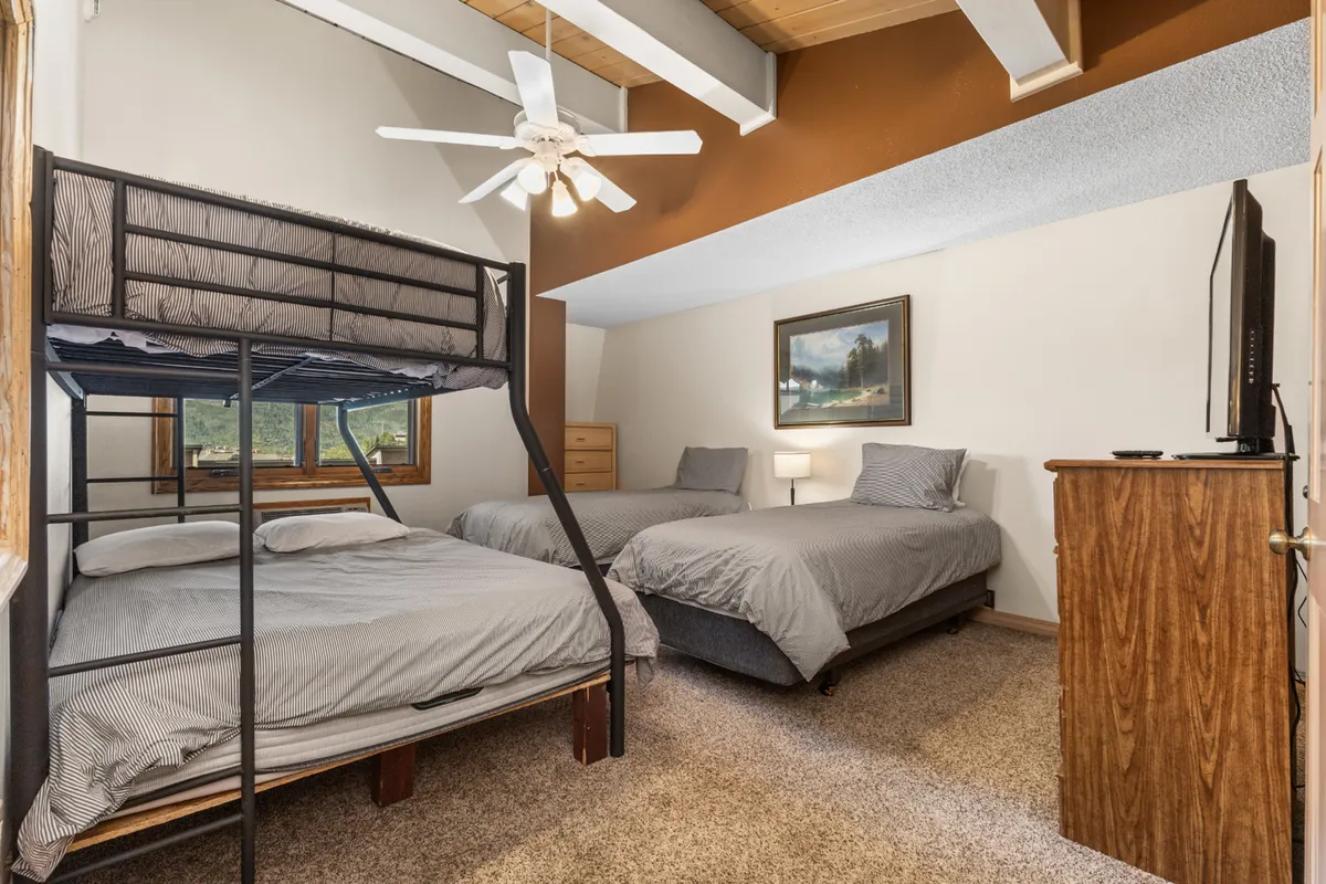 A cozy bedroom featuring a bunk bed and two double beds, complemented by a wooden dresser, framed landscape painting, and a ceiling fan. The room radiates a relaxed, comfortable mood suitable for families or groups with diverse sleeping needs.