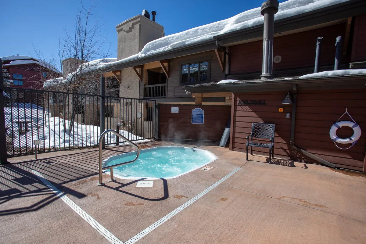 A cozy outdoor hot tub set in a fenced area, surrounded by snow. A lounge chair sits nearby, and the hot tub is steaming invitingly. The scene captures the pleasure of soaking in warmth while enjoying the crisp winter air, with snowy scenery adding to the sense of luxury and seasonal charm.