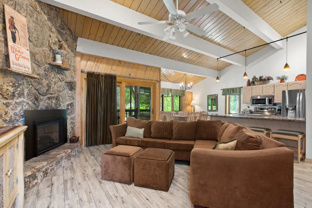 A spacious open-plan living area featuring a prominent stone fireplace, a large sectional sofa, and visible kitchen area. Large windows frame views of the outdoors, and the inviting setup is ideal for gatherings, conversation, and relaxation after outdoor adventures.