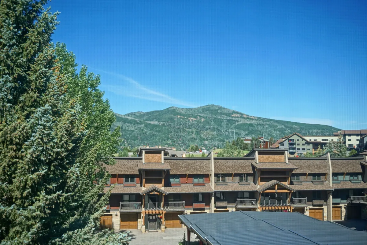 A picturesque exterior view of the condo building set against a stunning mountain backdrop and framed by tall trees. The scene radiates tranquility and highlights the breathtaking natural surroundings accessible right outside the door—ideal for hiking, biking, and exploration.