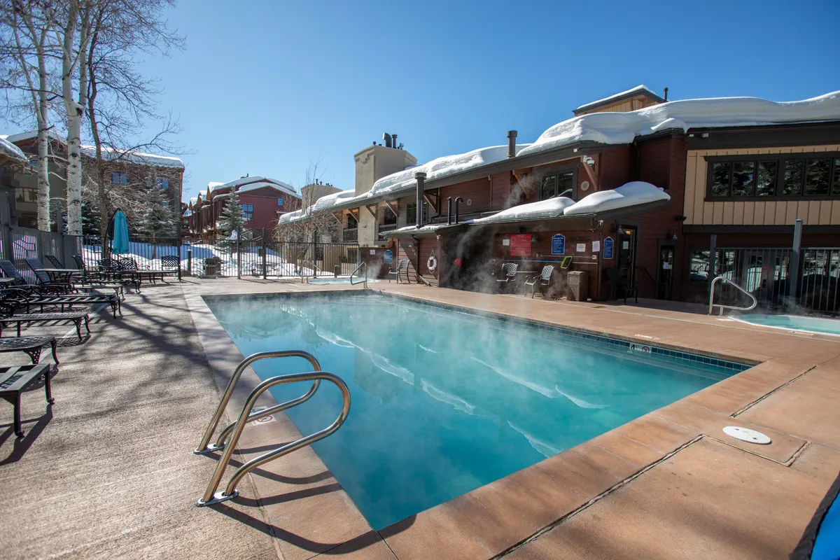 A steaming, blue swimming pool surrounded by snow, lounge chairs, and the resort building. The pool area feels rejuvenating and community-oriented, perfect for families or groups to enjoy après-ski relaxation together. The winter backdrop highlights the pool’s appeal even in colder months.