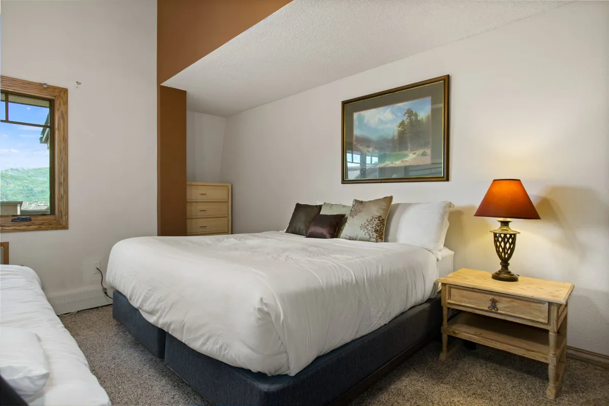 A spacious bedroom with a large, plush bed dressed in crisp white linens and decorative pillows, a nightstand with a lamp, a framed landscape painting, and a window revealing beautiful outdoor scenery. The space feels serene and private, emphasizing comfort and relaxation.