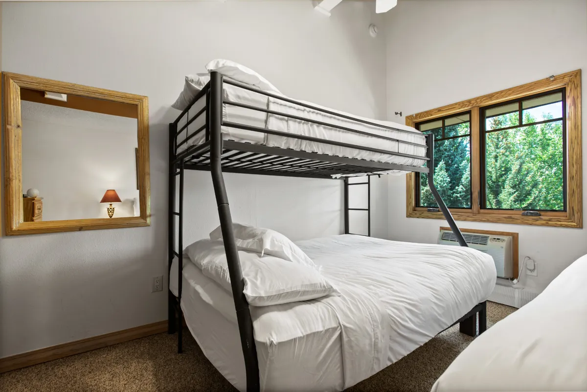 A cozy bedroom featuring a bunk bed and large window that lets in ample sunlight. The wooden mirror and air conditioning unit are visible, indicating comfort in both summer and winter. The bedding and overall layout create a welcoming, family-friendly environment ideal for relaxing after mountain adventures.