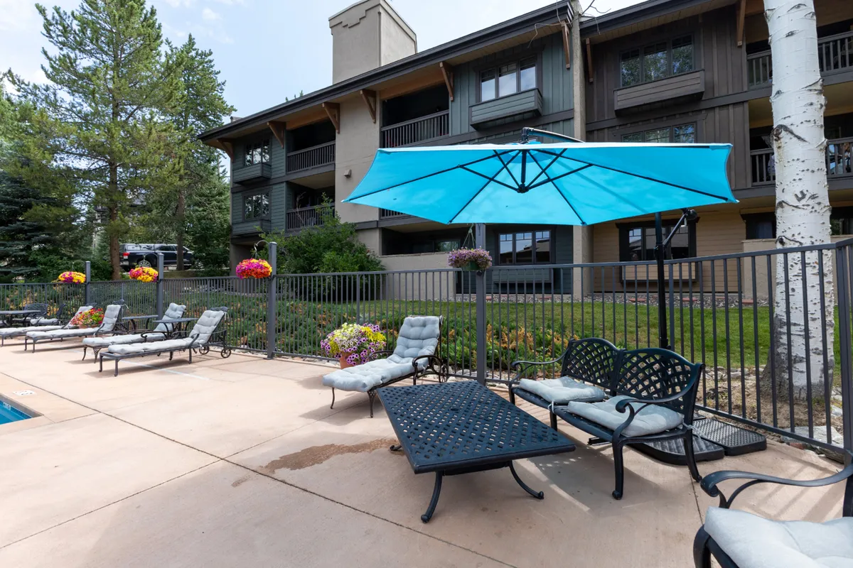 A bright exterior scene featuring a sparkling swimming pool surrounded by blue umbrellas, lounge chairs, and vibrant flower beds. The setup encourages relaxation and socializing, with the pool area serving as a centerpiece of the resort's amenities. The tranquil atmosphere is enhanced by lush landscaping and the inviting layout of the poolside seating.
